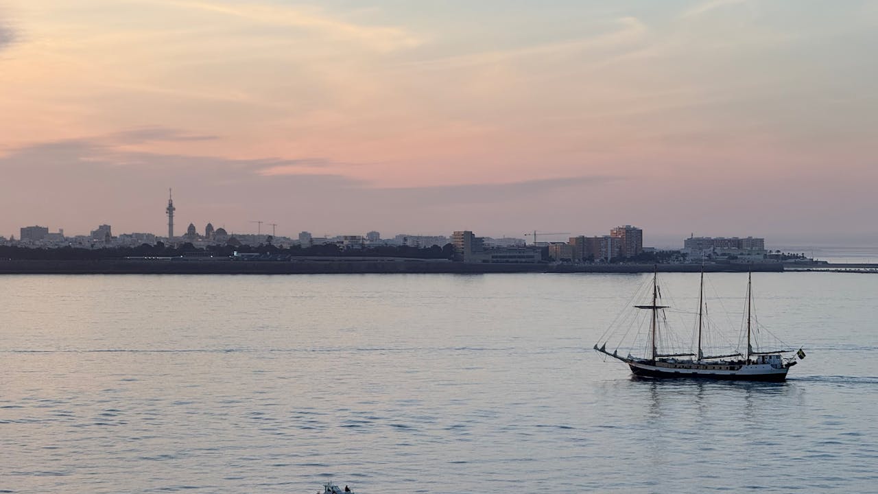 A picturesque view of a sailing ship at sunset with the Cádiz skyline in the background.